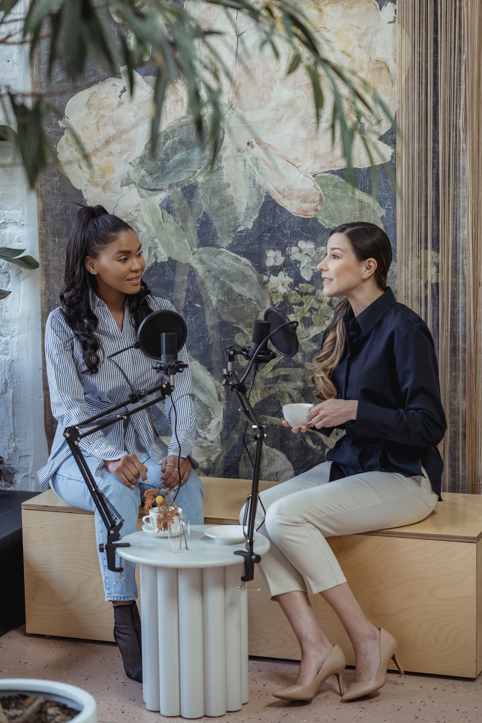 Two people talking into podcast microphones on boom arms, seated by a floral backdrop with coffee on a small table